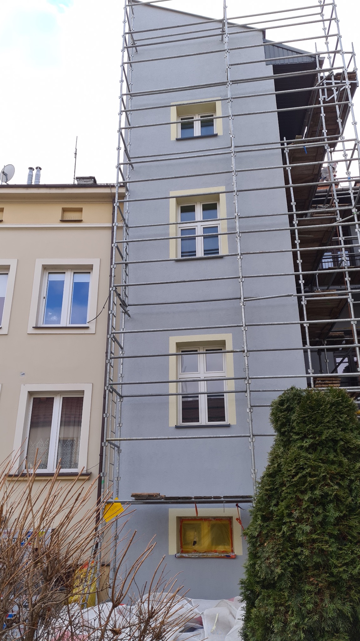 Szara elewacja budynku w trakcie prac, z widocznymi rusztowaniami, zabezpieczonym oknem folią i fragmentem sąsiedniej, beżowej elewacji.