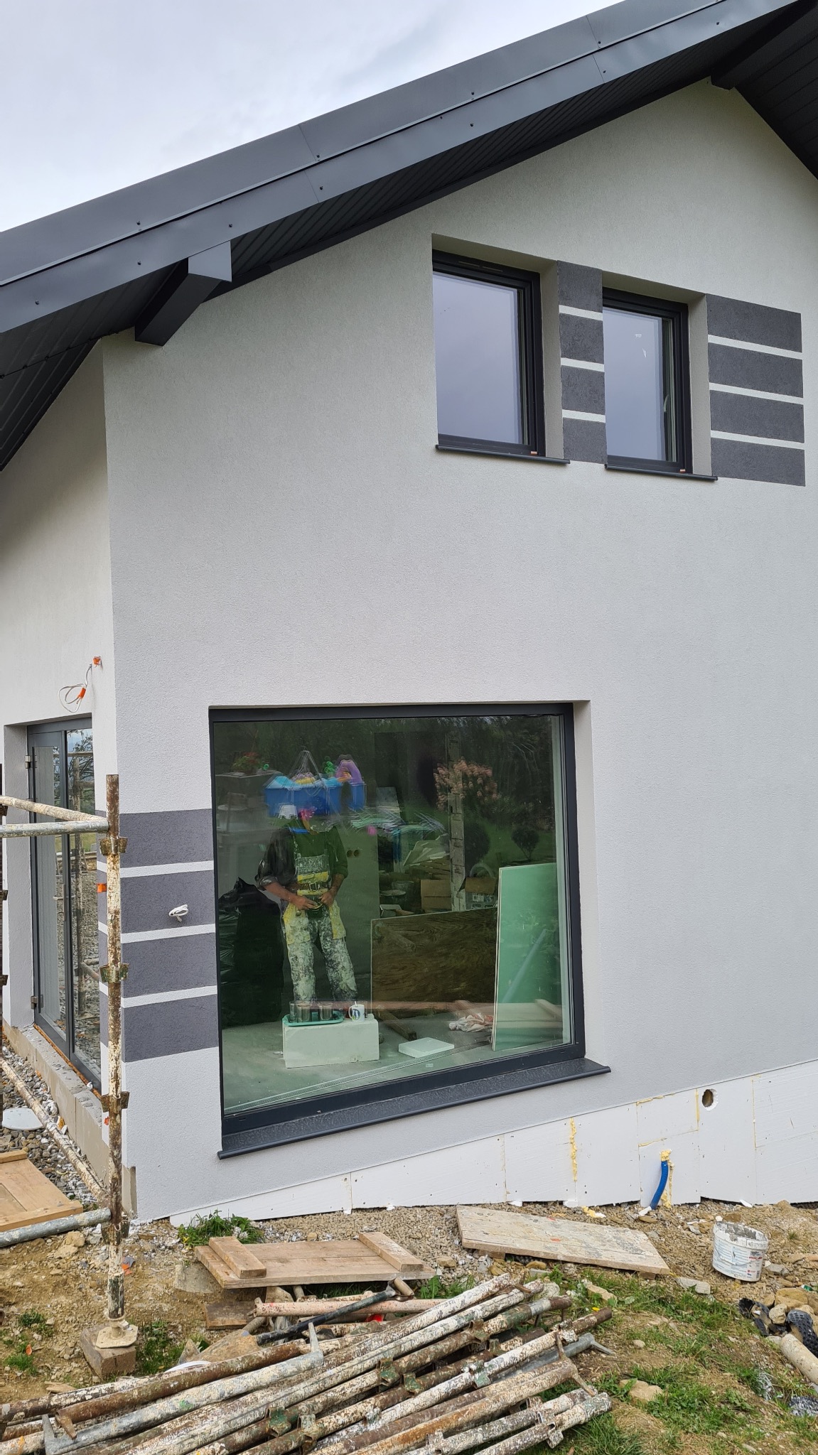 Nowoczesny dom z szarą elewacją i dekoracyjnymi pasami, widoczne rusztowania i odbicie pracownika w oknie.