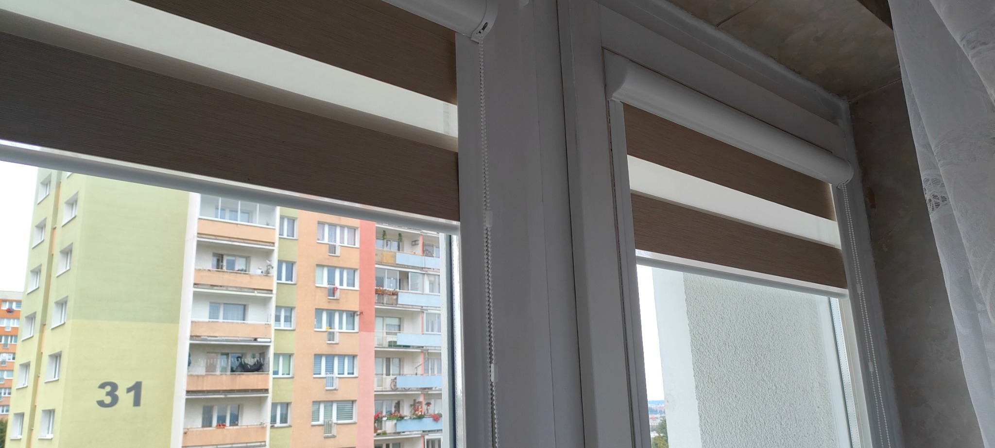 Zamknięta roleta dzień noc w oknie z widokiem na blok mieszkalny. Widoczne poziome pasy rolety w odcieniach brązu i bieli. Szary budynek za oknem.