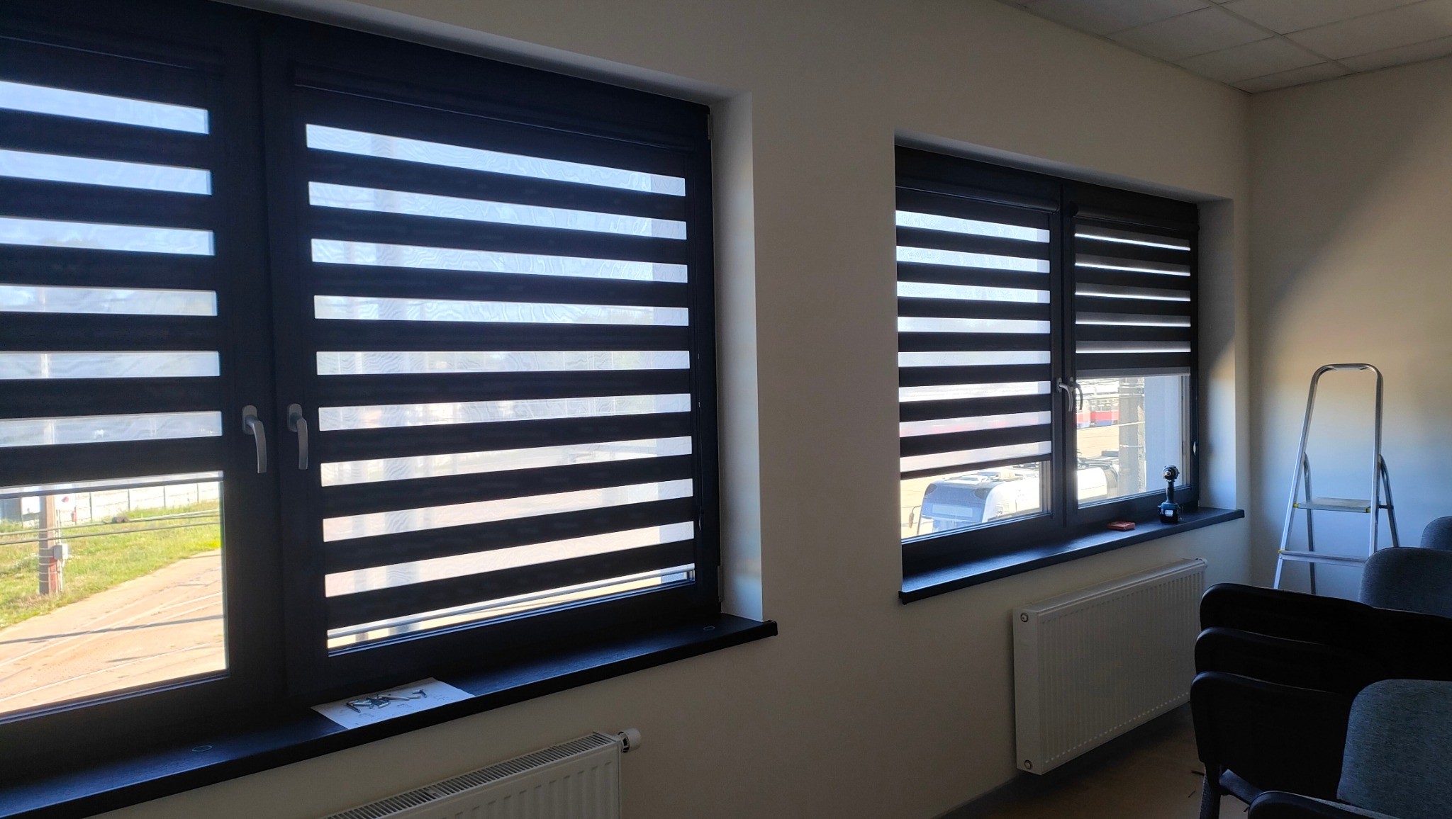 Dwa okna z zamontowanymi roletami dzień noc w kolorze antracytowym. Widok na zewnątrz, gdzie widać budynek MZK. Na parapecie leży kartka i wkrętarka.