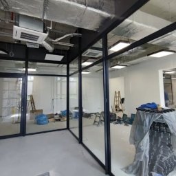 NOVAOKNA KM SPÓŁKA Z OGRANICZONĄ ODPOWIEDZIALNOŚCIĄ - Montaż szklanych ścian działowych w biurze, widoczne narzędzia i drabiny, surowy stan wykończenia, instalacja klimatyzacji na suficie.