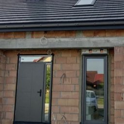 NOVAOKNA KM SPÓŁKA Z OGRANICZONĄ ODPOWIEDZIALNOŚCIĄ - Szare drzwi wejściowe i okno z roletą zewnętrzną w surowym stanie budynku z cegły, widoczne przewody i paleta budowlana.