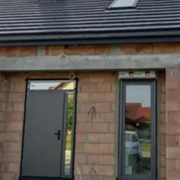 Szare drzwi wejściowe i okno z roletą zewnętrzną w surowym stanie budynku z cegły, widoczne przewody i paleta budowlana.