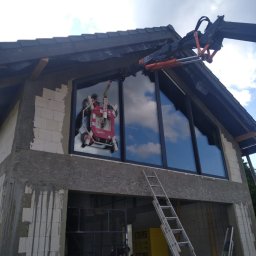 NOVAOKNA KM SPÓŁKA Z OGRANICZONĄ ODPOWIEDZIALNOŚCIĄ - Montaż okna na elewacji domu w stanie surowym za pomocą specjalistycznego podnośnika. Widoczne drabiny i materiały budowlane.