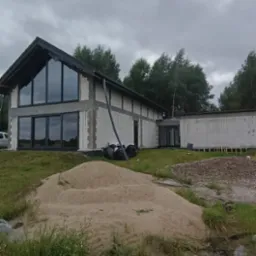 Dom w trakcie budowy z dużymi oknami, widoczne bloczki i piasek. Czarna stolarka okienna, pochmurne niebo w tle.