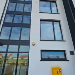 Elewacja budynku z oknami o czarnych ramach, w tym duże okno fasadowe. Żółta skrzynka gazowa przy ścianie. Budynek o nowoczesnej architekturze.