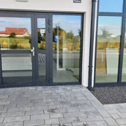 Szklane drzwi wejściowe z szarymi ramami, widok z zewnątrz na budynek mieszkalny i otoczenie. Na drzwiach widoczna tabliczka informacyjna.