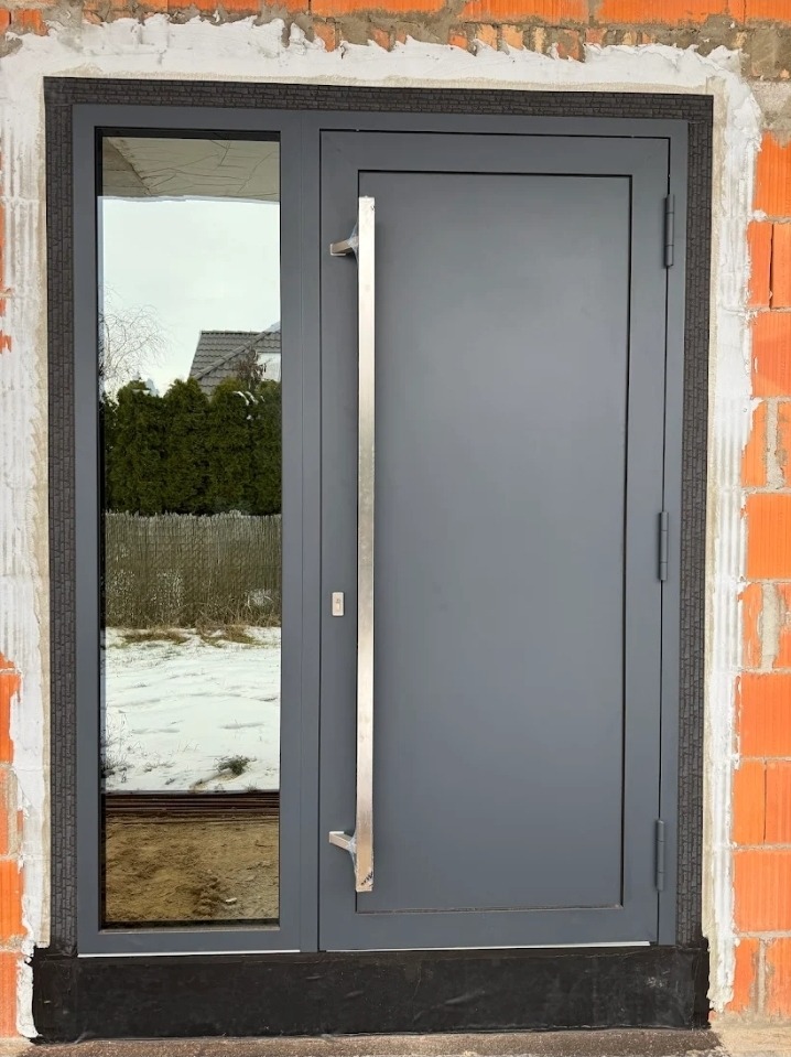 Szare drzwi wejściowe z długim, srebrnym uchwytem i szybą boczną, zamontowane w surowym murze z cegły. Widok na ośnieżony ogród.