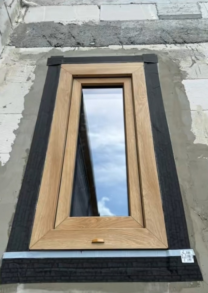 Drewniane okno uchylne z widocznym montażem w surowej ścianie, Bytów. Detal wykończenia i uszczelnienia na tle nieba odbitego w szybie.