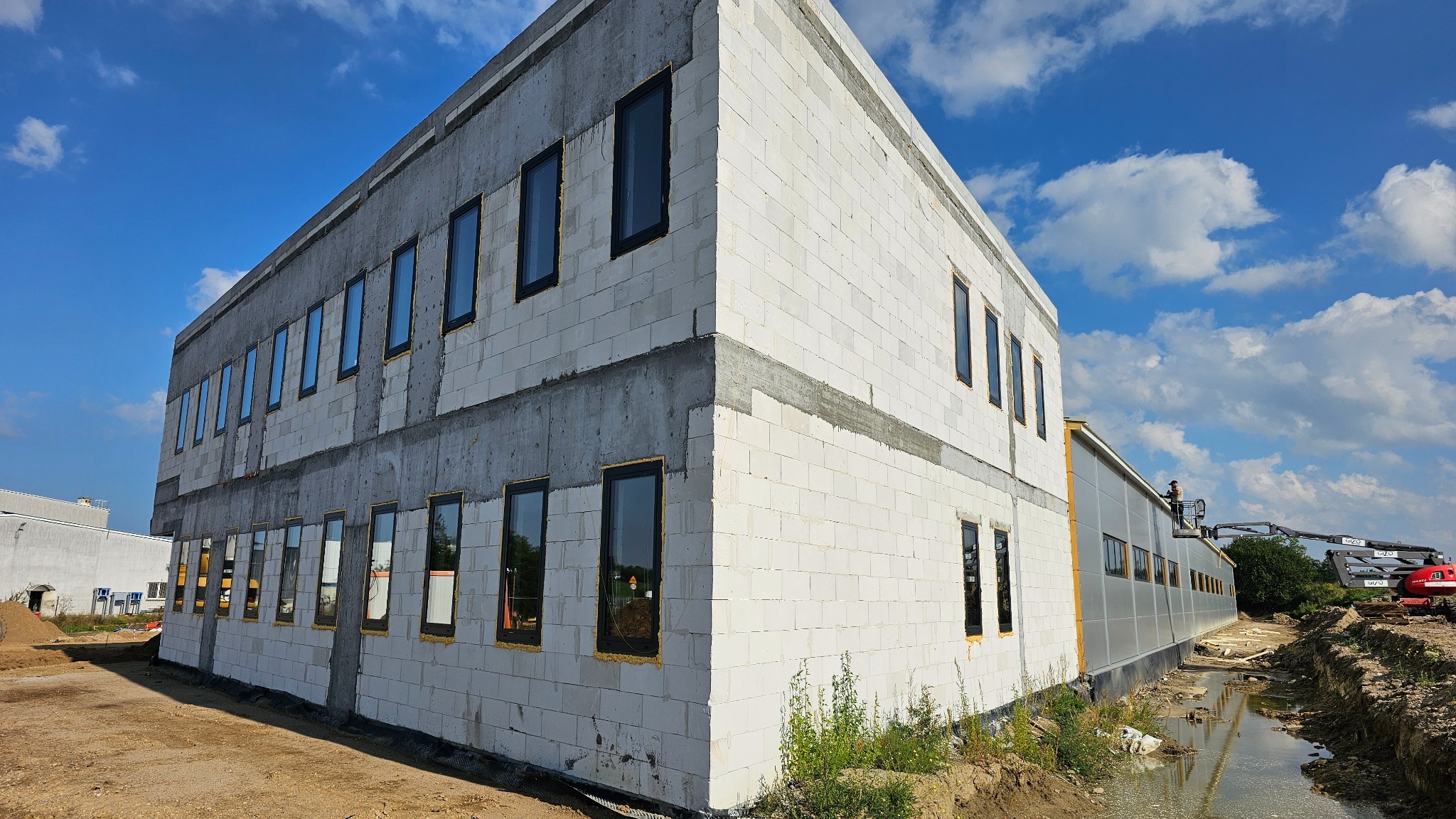 Budynek w trakcie budowy: białe bloczki, czarne okna, widoczne ślady zaprawy. Niebo z chmurami w tle. Po prawej stronie budynku widoczny podnośnik koszowy.