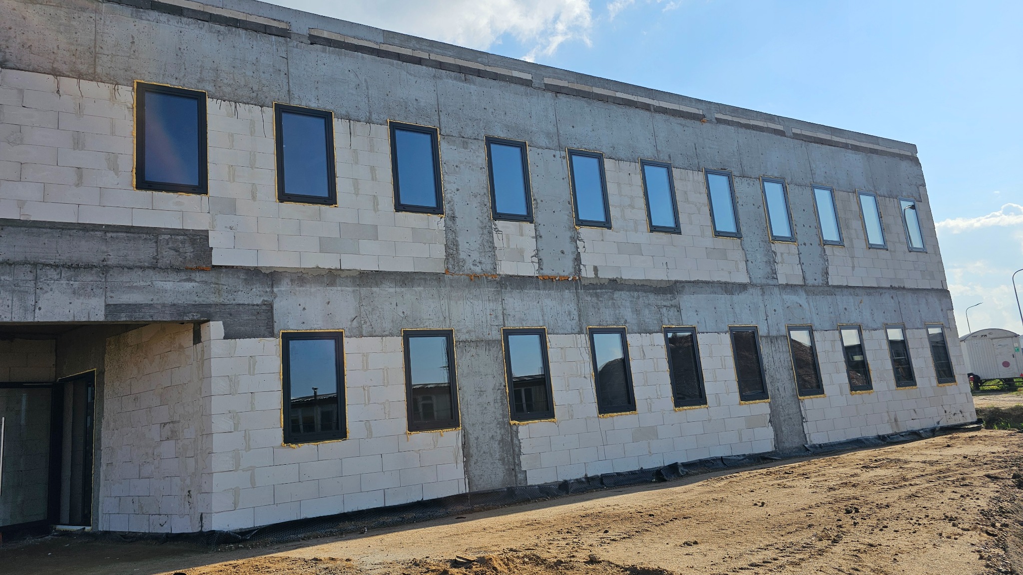 Nowo wstawione okna w surowym budynku z betonu komórkowego, widoczne ślady pianki montażowej. Część okien z odbiciami nieba, inne ciemne. Budynek w trakcie budowy.