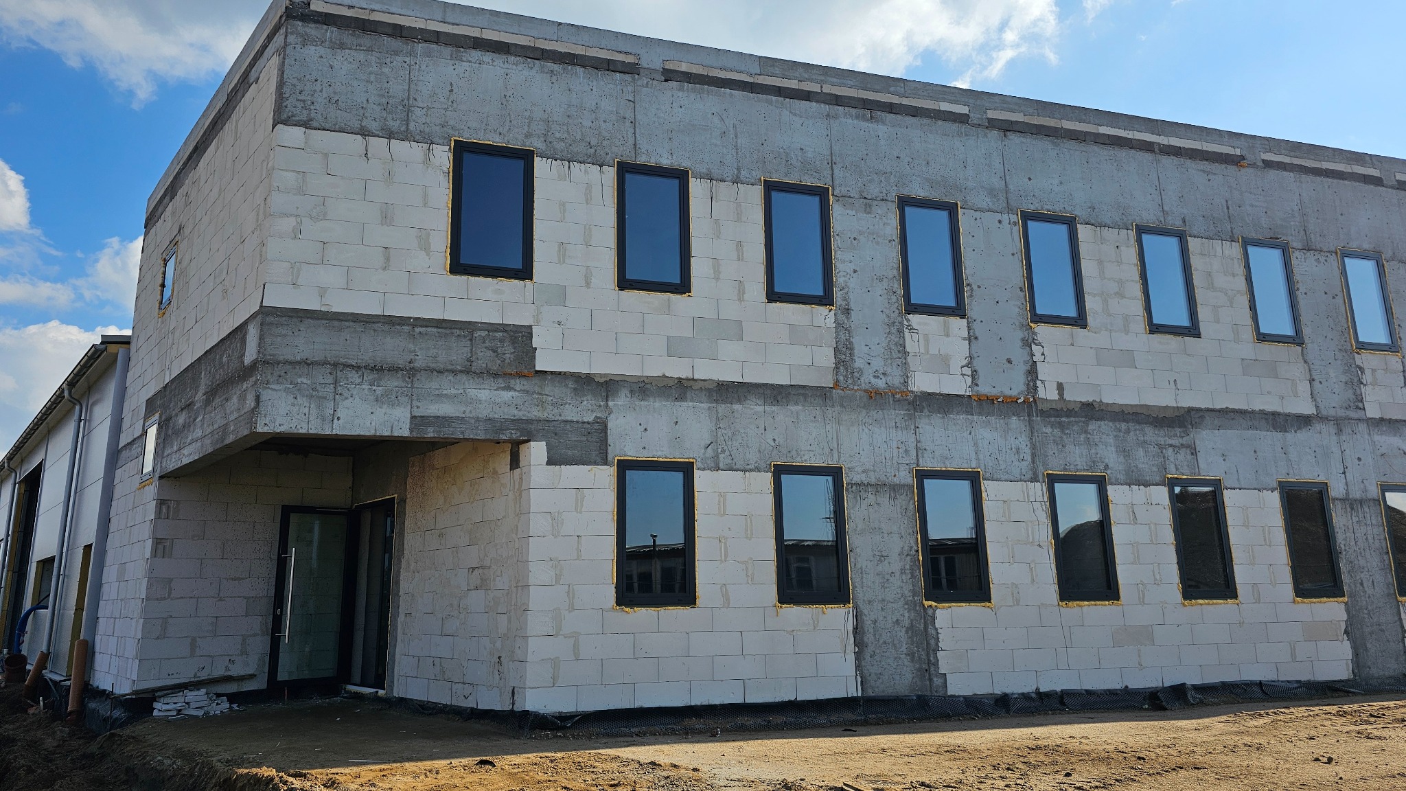 Budynek w trakcie budowy z zamontowanymi oknami z czarną ramą. Widoczne bloczki betonowe i wstępne wykończenie elewacji. Błękitne niebo w tle.