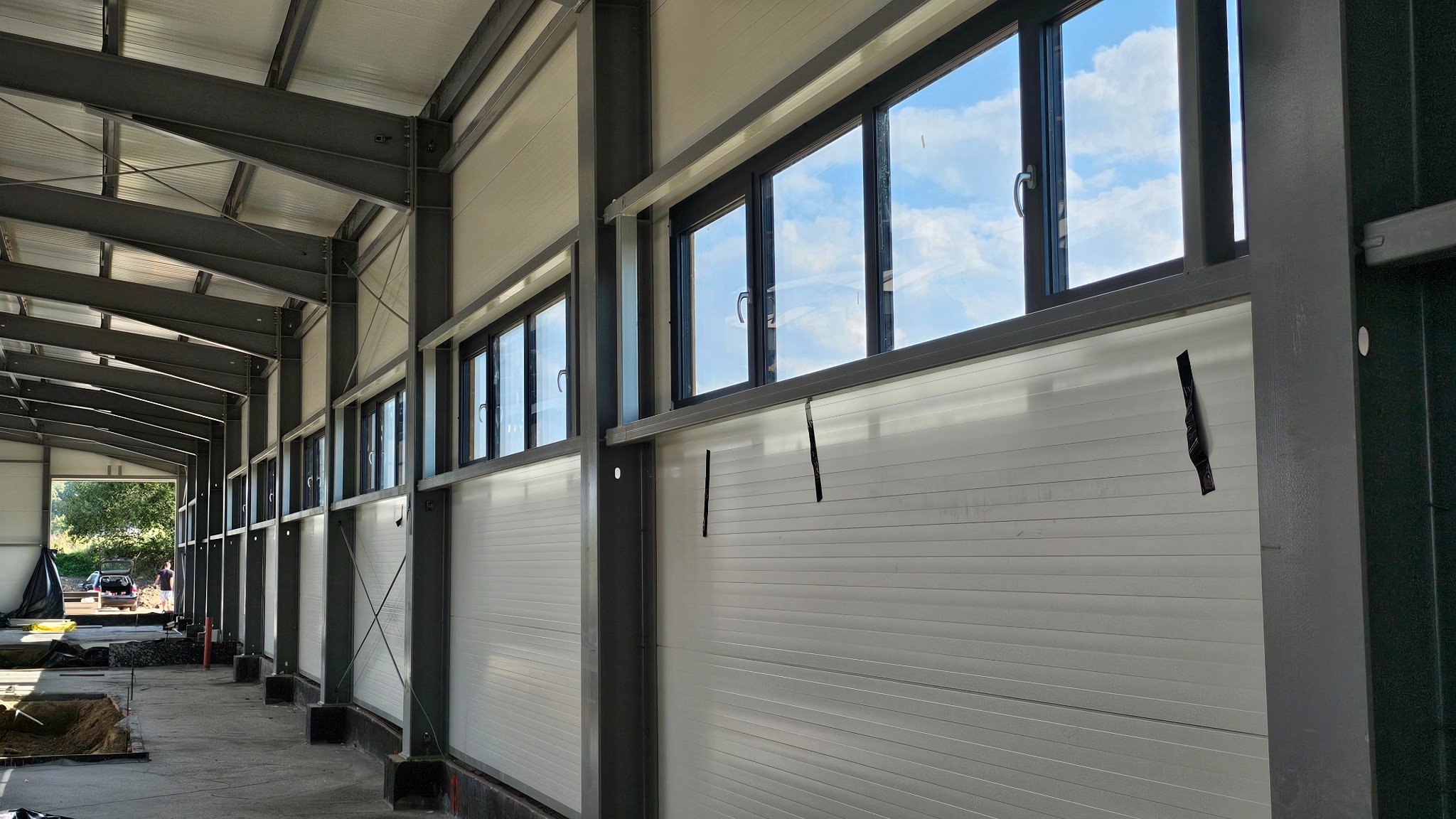 Wnętrze hali produkcyjnej w trakcie budowy. Widoczne okna w metalowych ramach, białe ściany z paneli i elementy konstrukcji stalowej. W tle widoczne otoczenie zewnętrzne.