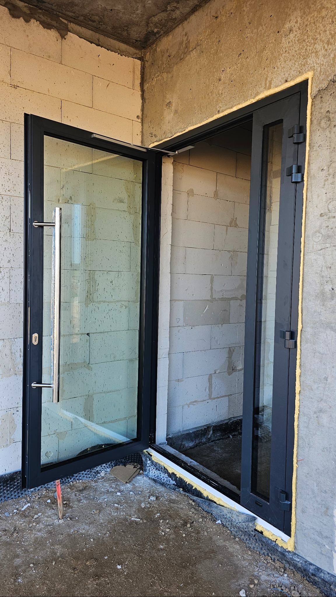 Czarne drzwi wejściowe z długim, srebrnym uchwytem, zamontowane w surowym stanie budynku. Widoczne bloczki i pianka montażowa.