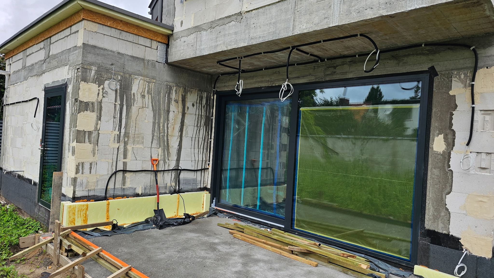 Montaż dużych, przesuwnych drzwi w surowym stanie budynku. Widoczne ocieplenie, kable i narzędzia. Dwa skrzydła drzwiowe zabezpieczone folią ochronną.