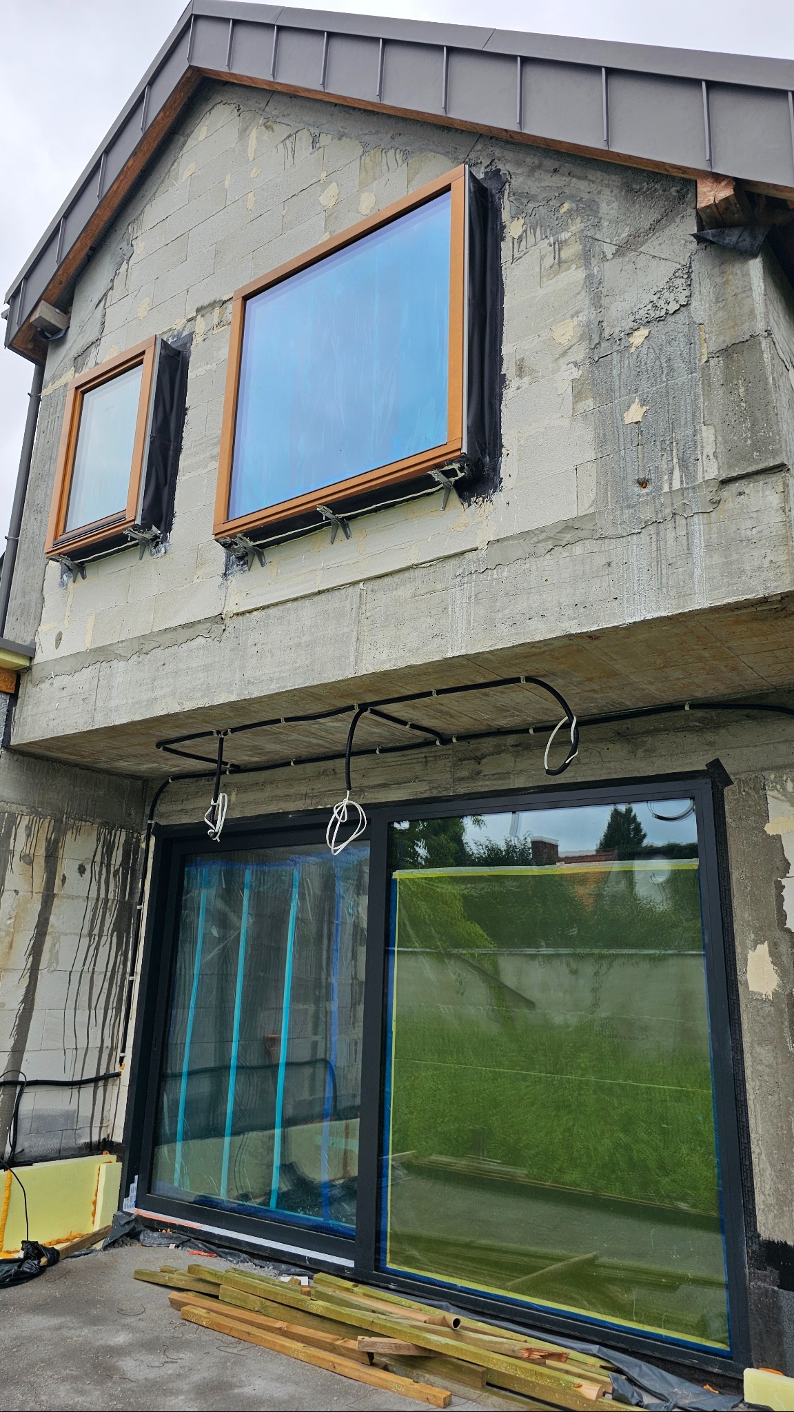 Montaż okien drewnianych w surowym budynku. Widoczne okna z drewnianymi ramami, instalacja elektryczna i materiały budowlane. Okna zabezpieczone folią.