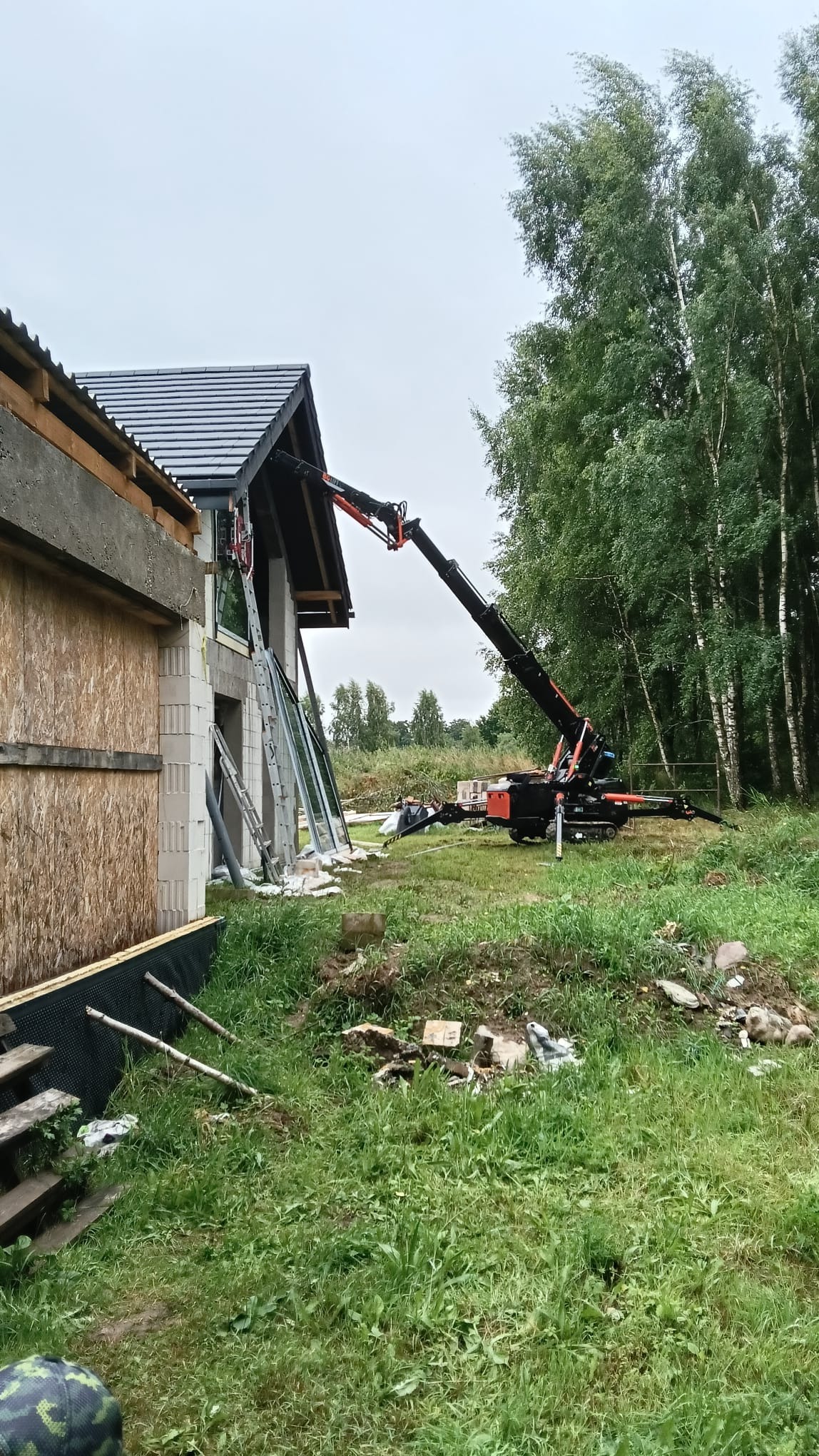 Montaż okna z użyciem dźwigu na budowie domu. Okna o dużych przeszkleniach. Widoczne materiały budowlane i zieleń. Pochmurne niebo.