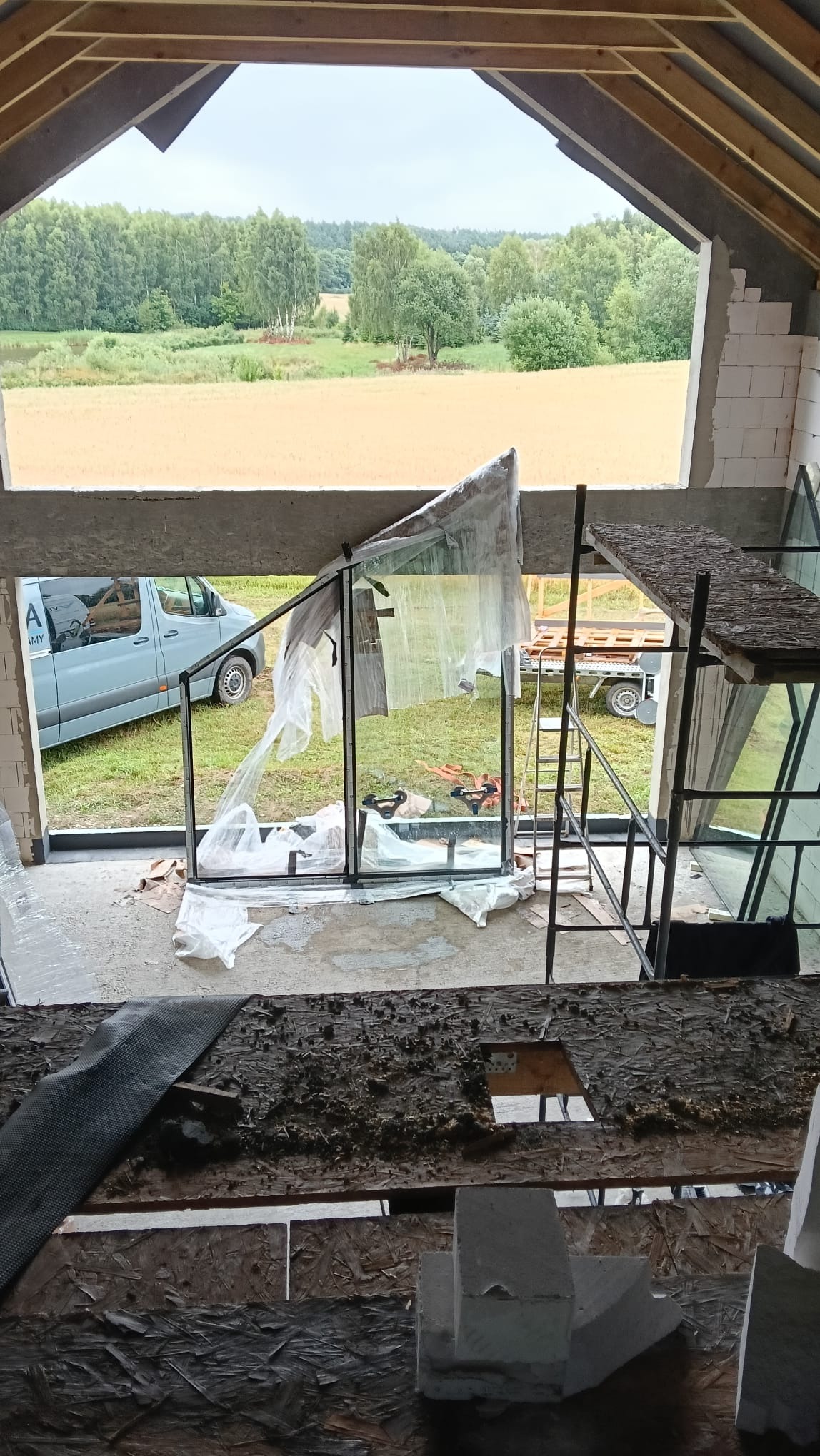 Montaż okna tarasowego z czarnymi ramami w surowym stanie. Widok na krajobraz za oknem, częściowo zakryte folią ochronną, rusztowanie obok.