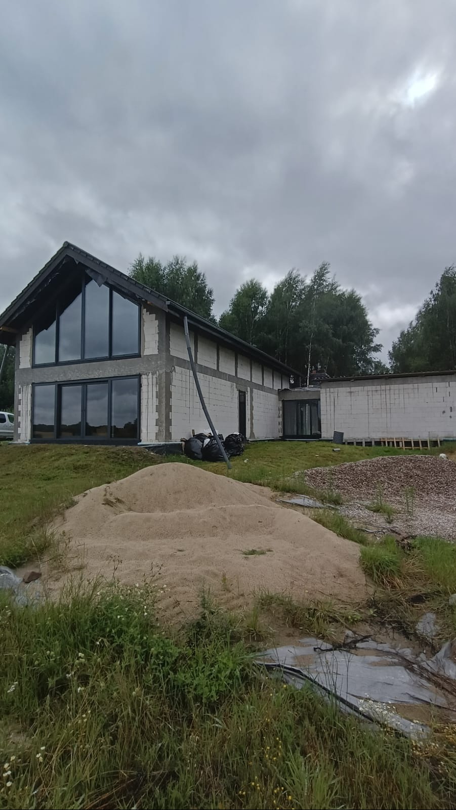 Dom w trakcie budowy z dużymi oknami, widoczne bloczki i piasek. Czarna stolarka okienna, pochmurne niebo w tle.