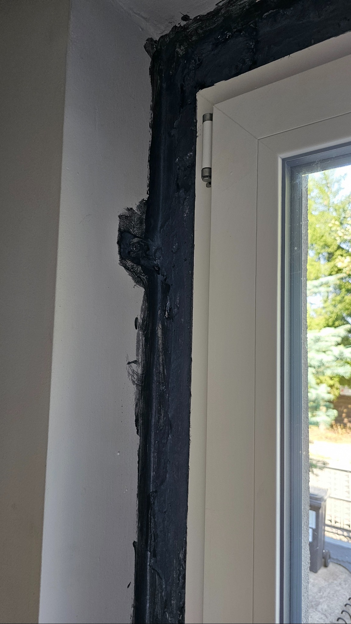Uszczelnianie ramy okiennej czarnym silikonem na styku ze ścianą, widoczne niedoskonałości i nierówności w nałożeniu materiału. Widok na zewnątrz przez okno.