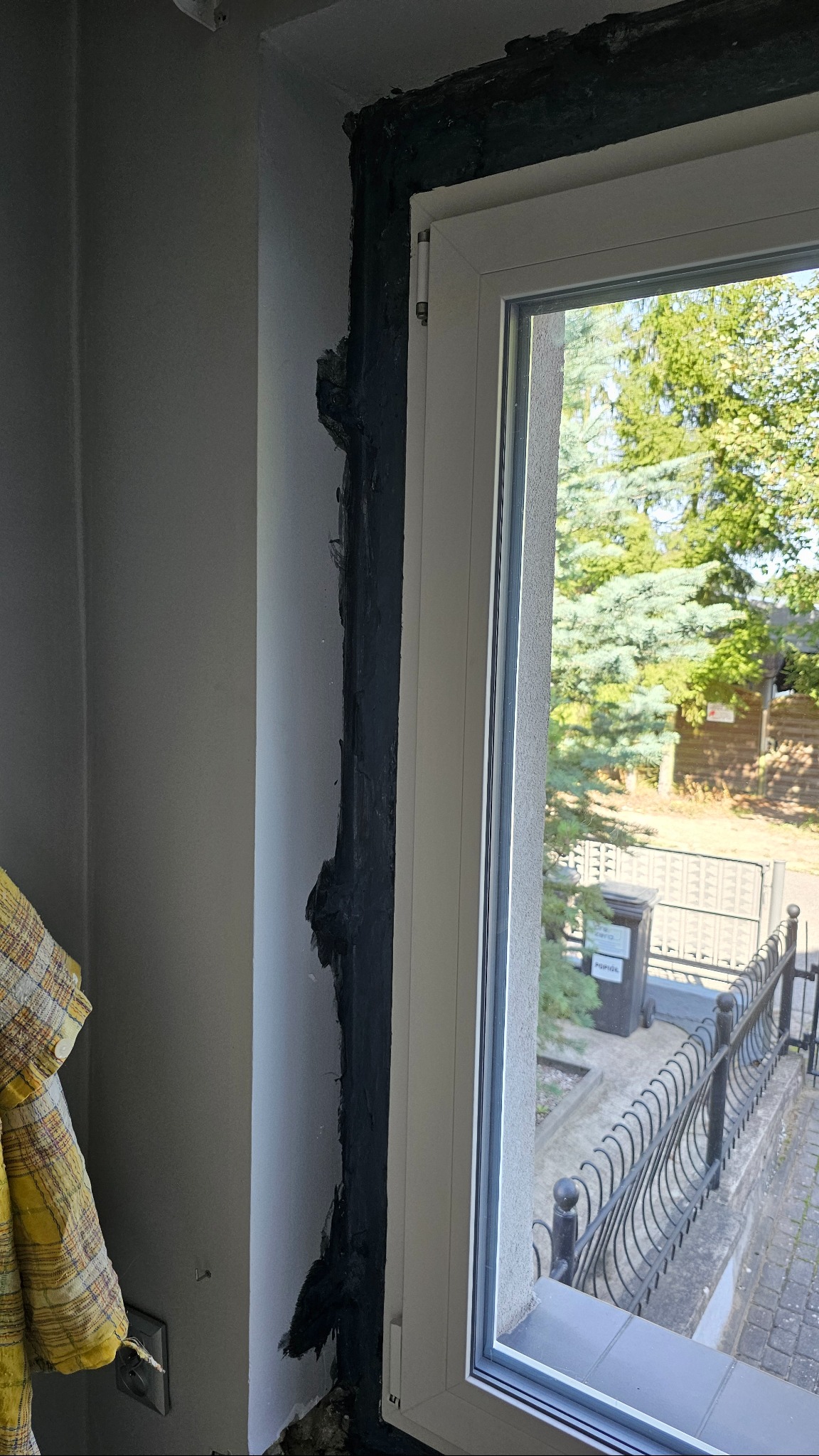 Białe okno PCV w trakcie montażu, widoczne czarne uszczelnienie i surowa ściana w tle. Widok na zieleń i ogrodzenie przez szybę.