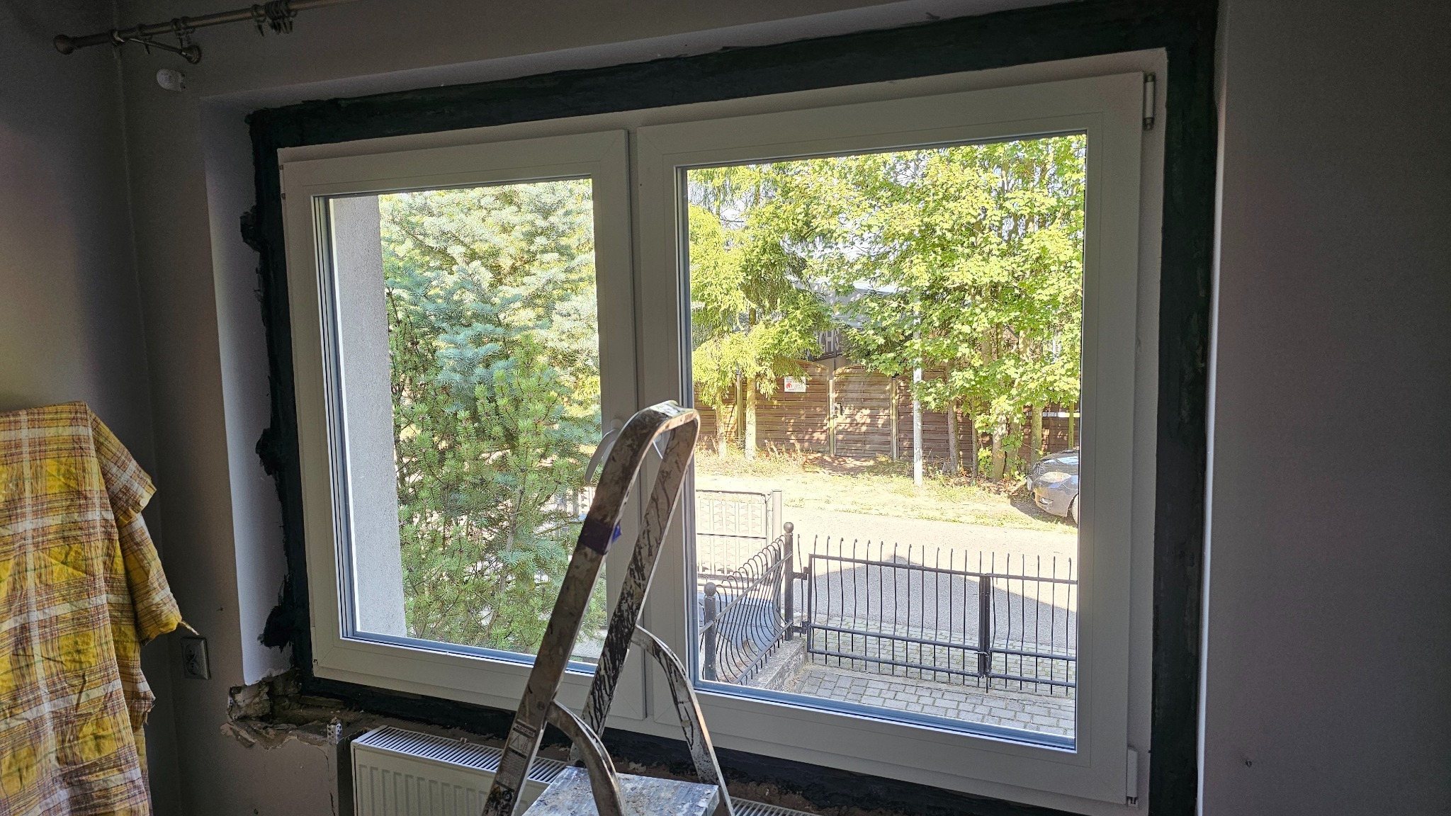 Montaż okna PCV: widok z wnętrza na białe okno z widokiem na zieleń, drabina oparta o parapet, czarne uszczelnienie ramy. Etap prac wykończeniowych.