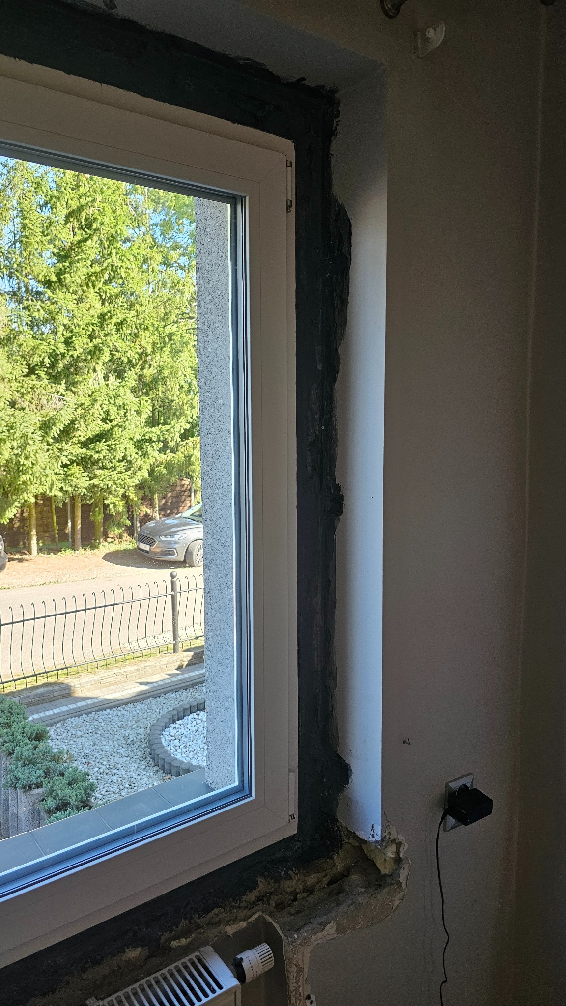 Białe okno PCV w trakcie montażu, widoczne uszczelnianie ramy czarną pianką montażową. Na zewnątrz widok na ogród i zaparkowany samochód.