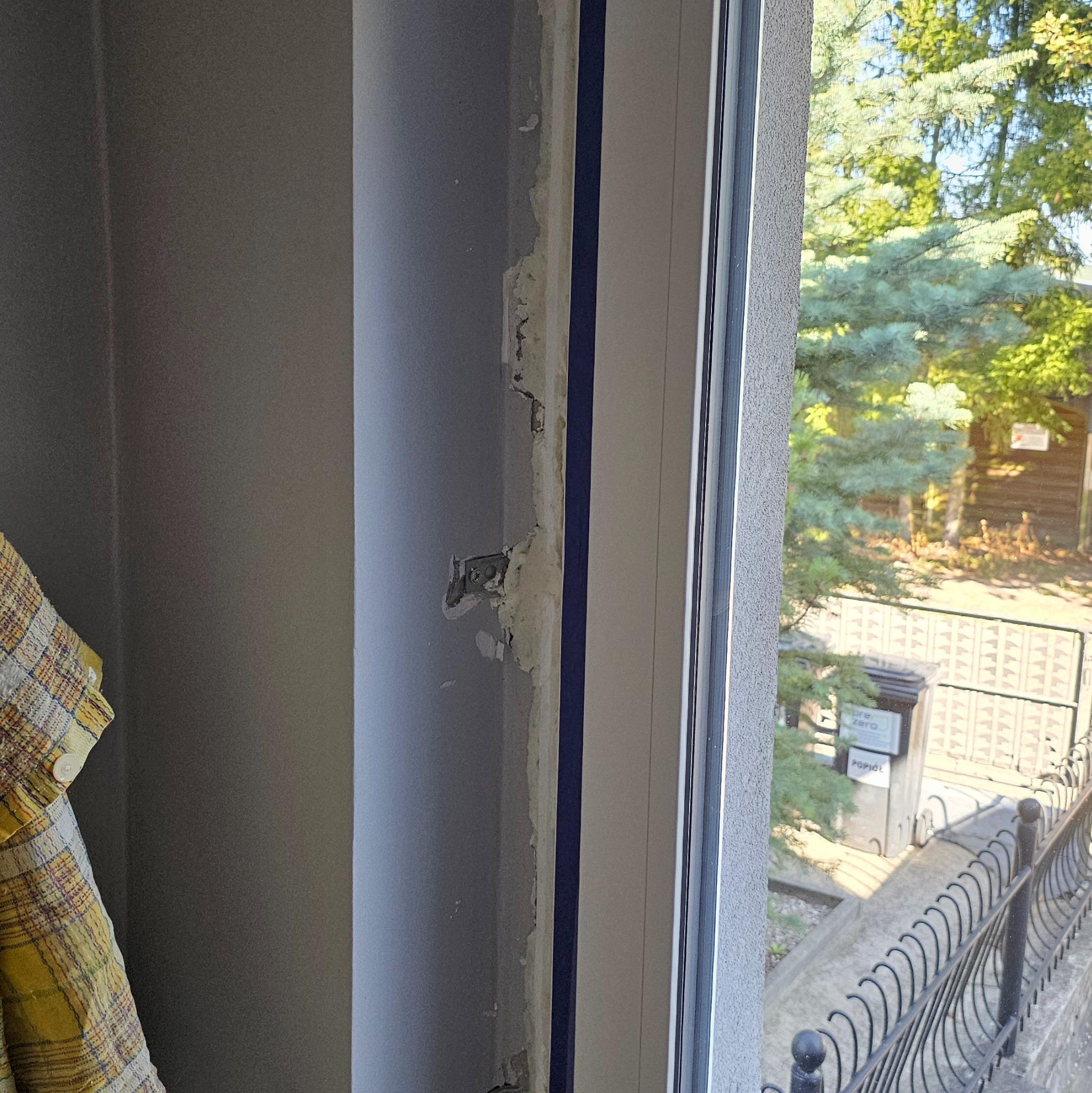 Fragment ściany z uszkodzonym tynkiem wokół ramy okiennej w trakcie montażu. Widoczne elementy mocujące i fragment widoku z okna na zewnątrz.