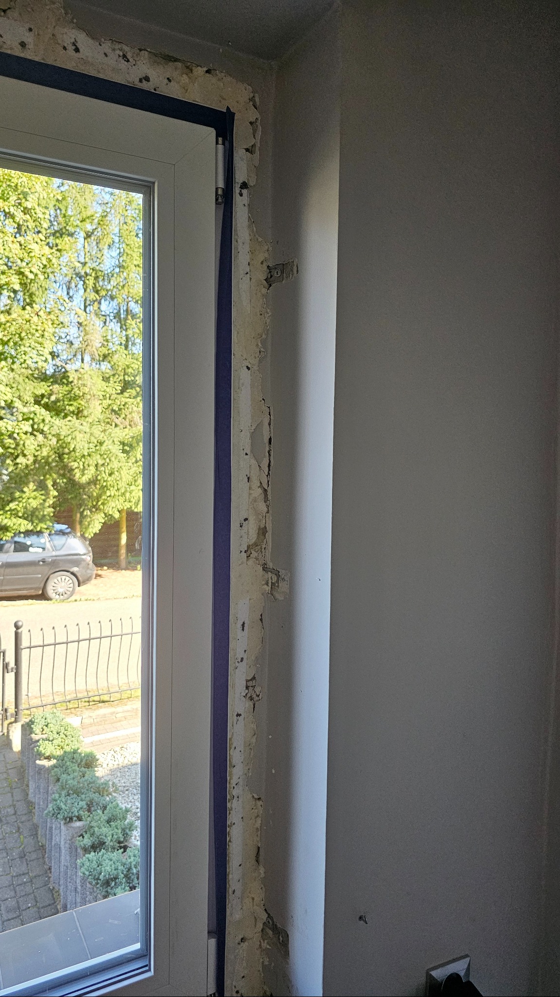 Białe okno PCV w trakcie montażu. Widoczne uszkodzenia ściany i taśma zabezpieczająca. Na zewnątrz zieleń i szary samochód. Wnętrze w trakcie prac wykończeniowych.