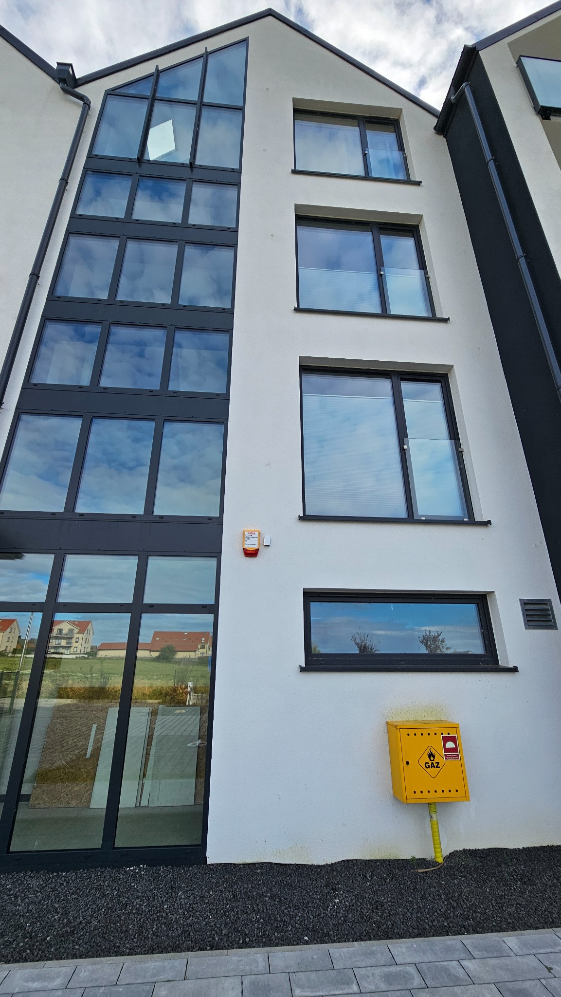 Elewacja budynku z oknami o czarnych ramach, w tym duże okno fasadowe. Żółta skrzynka gazowa przy ścianie. Budynek o nowoczesnej architekturze.