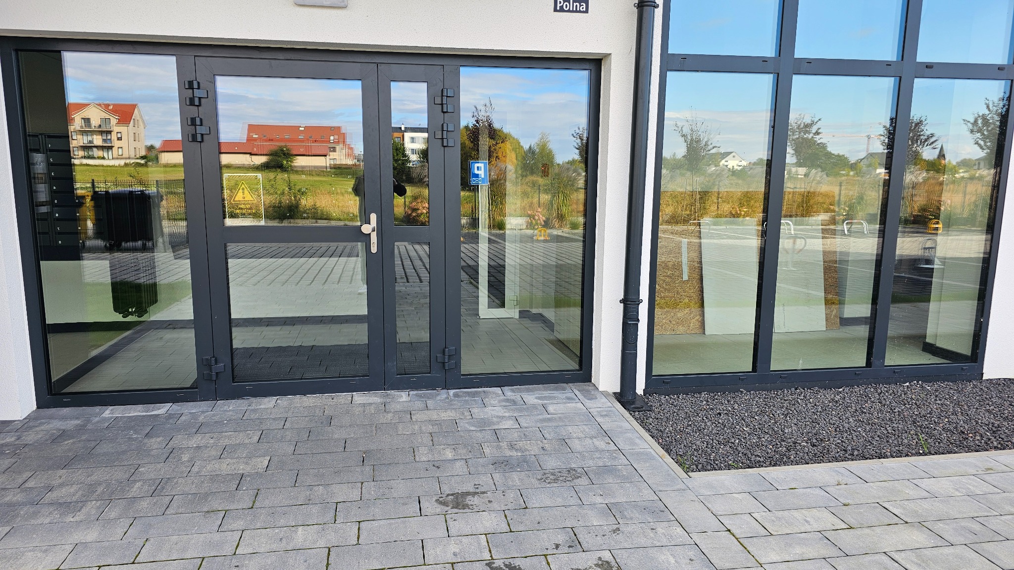 Szklane drzwi wejściowe z szarymi ramami, widok z zewnątrz na budynek mieszkalny i otoczenie. Na drzwiach widoczna tabliczka informacyjna.