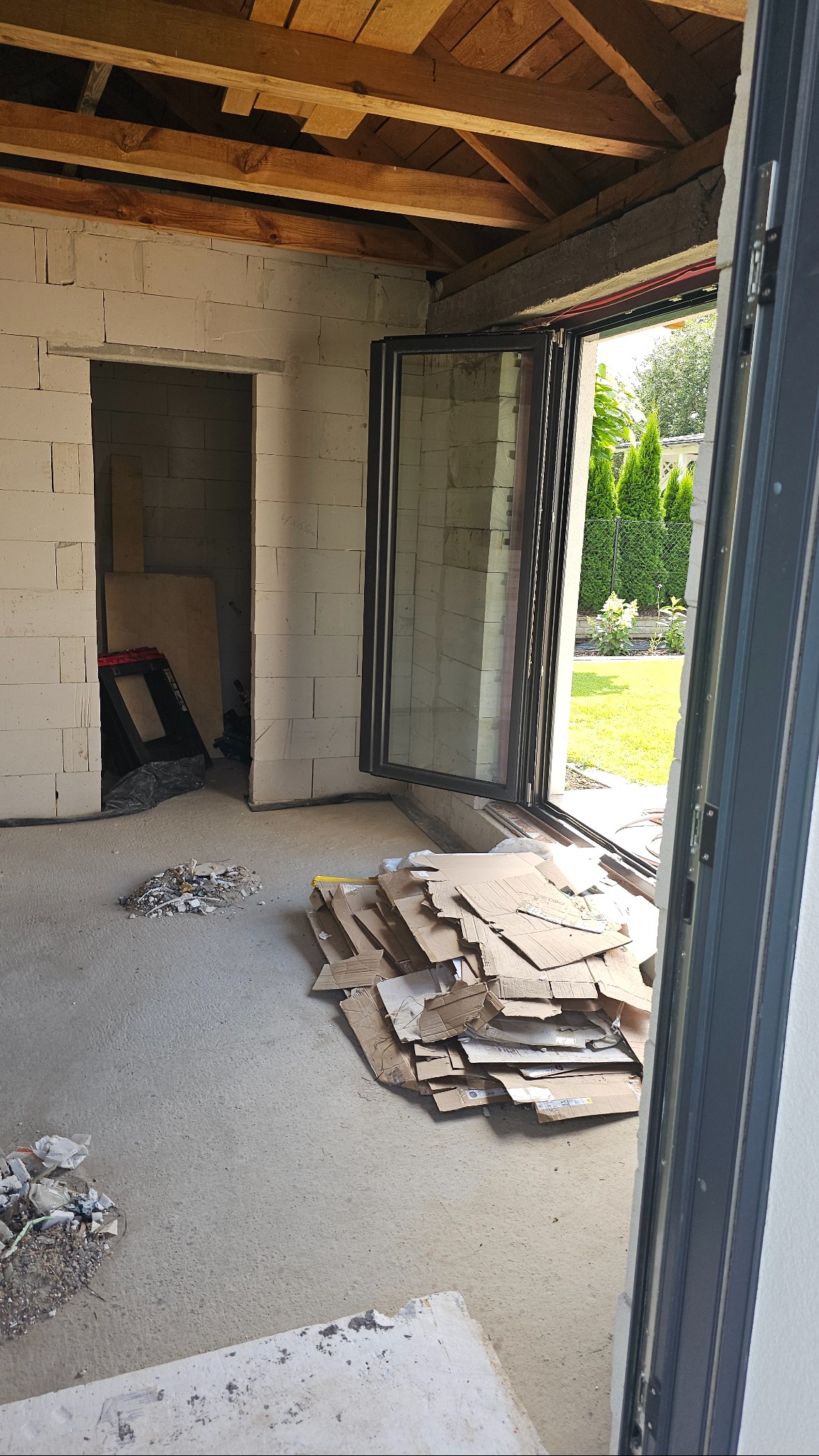 Montaż okna aluminiowego w surowym wnętrzu. Widok na ogród przez otwarte okno, w tle sterta kartonów i materiałów budowlanych.