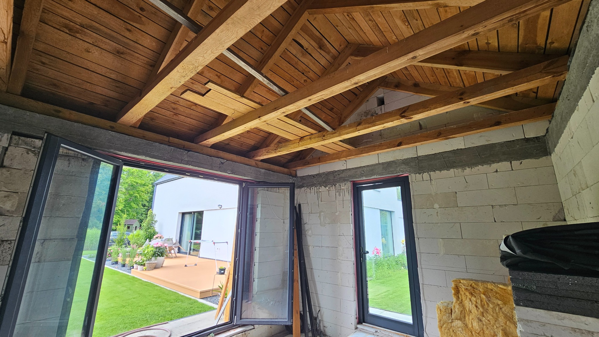 Surowy stan: czarne ramy okienne, ściany z betonu komórkowego i drewniany strop w nowym budynku z widokiem na ogród i taras.