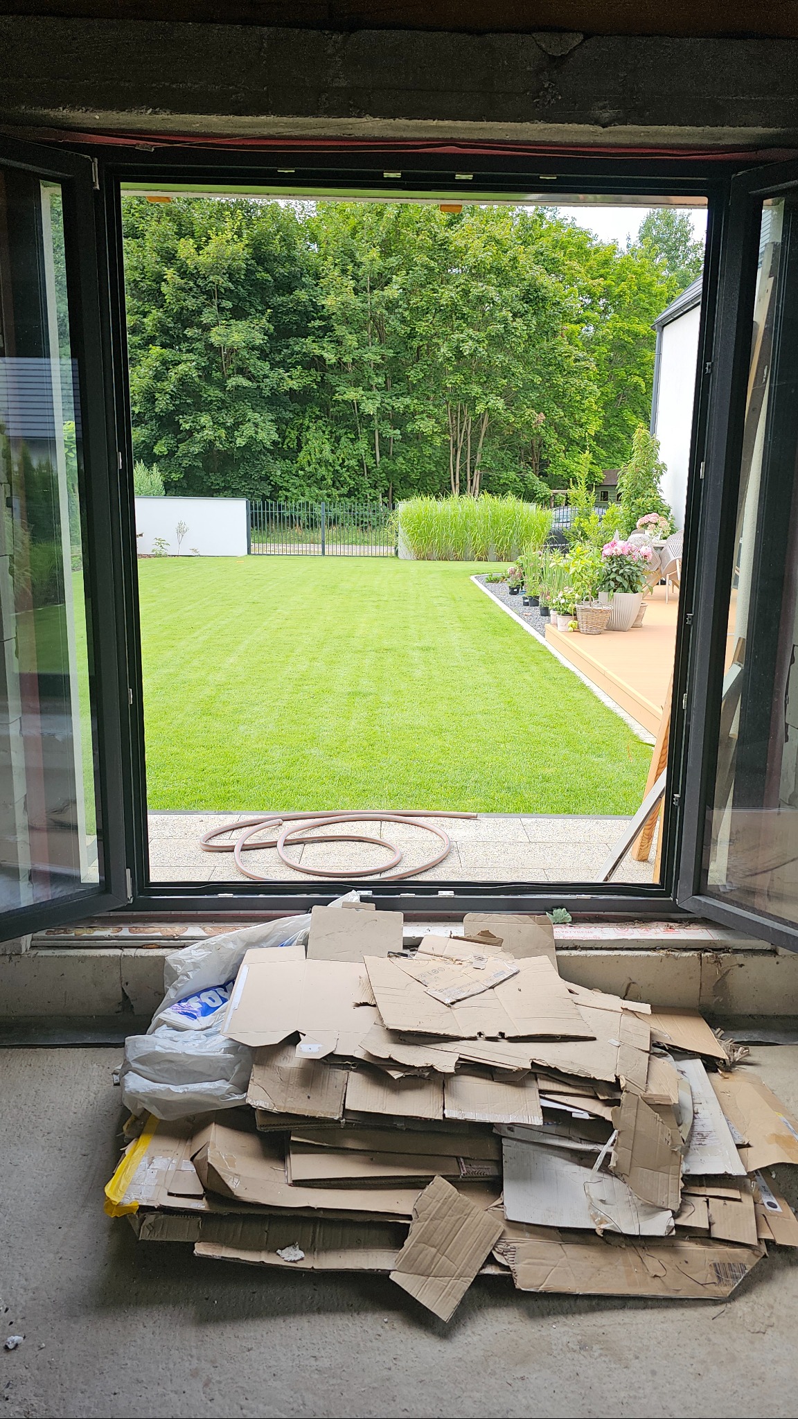 Otwarty, czarny system przesuwny z widokiem na zadbany ogród. Na pierwszym planie kartony i folie, co sugeruje prace wykończeniowe.