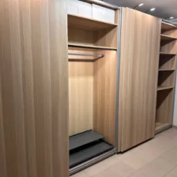 Jasna, drewniana szafa wnękowa z przesuwnymi drzwiami, oświetleniem punktowym i częściowo wysuniętymi półkami na buty, prezentująca funkcjonalne wnętrze.