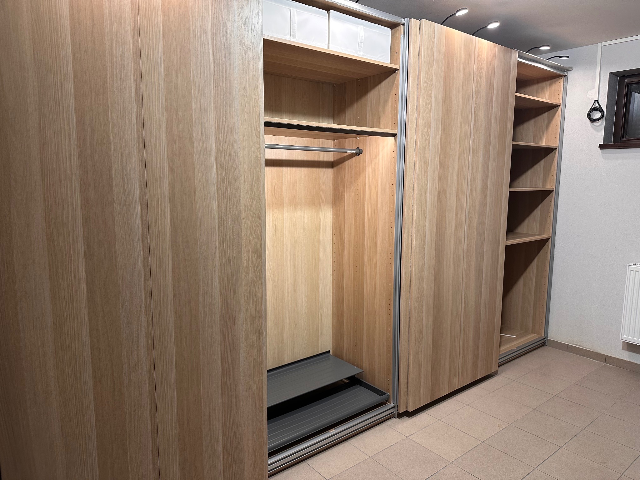 Jasna, drewniana szafa wnękowa z przesuwnymi drzwiami, oświetleniem punktowym i częściowo wysuniętymi półkami na buty, prezentująca funkcjonalne wnętrze.