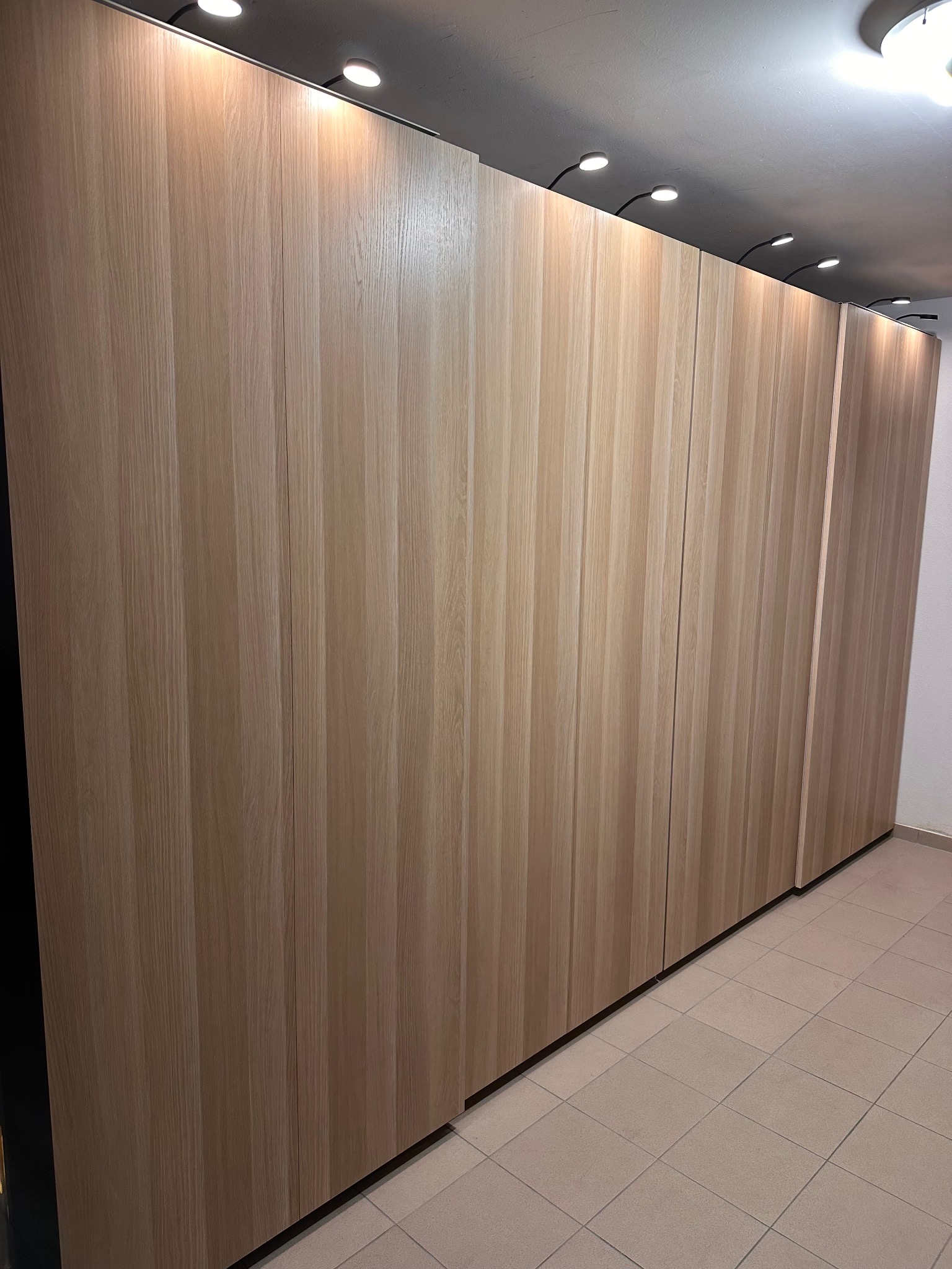 Długa, zabudowana szafa wnękowa z jasnego drewna, oświetlona punktowymi lampami sufitowymi. Widoczne pionowe panele drzwiowe i płytki podłogowe.