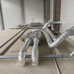 OLIMA SPÓŁKA Z OGRANICZONĄ ODPOWIEDZIALNOŚCIĄ - Rozbudowany system kanałów wentylacyjnych z izolacją aluminiową w surowym wnętrzu hali, widoczne dwa centrale wentylacyjne, drabina w tle, szare ściany i betonowa podłoga.