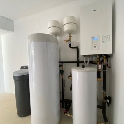 OLIMA SPÓŁKA Z OGRANICZONĄ ODPOWIEDZIALNOŚCIĄ - Nowoczesny, biały piec gazowy Panasonic Aquarea Smart Cloud z podłączonymi zbiornikami, rurami i zaworami w minimalistycznym, białym pomieszczeniu; widoczny zmiękczacz wody z boku.