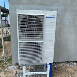 OLIMA SPÓŁKA Z OGRANICZONĄ ODPOWIEDZIALNOŚCIĄ - Biała jednostka zewnętrzna pompy ciepła marki Panasonic zamontowana na białym stelażu przy szarej ścianie budynku w trakcie budowy, z widocznymi niebieskimi rurami i rusztowaniem w tle.