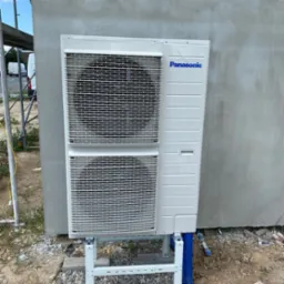 Biała jednostka zewnętrzna pompy ciepła marki Panasonic zamontowana na białym stelażu przy szarej ścianie budynku w trakcie budowy, z widocznymi niebieskimi rurami i rusztowaniem w tle.