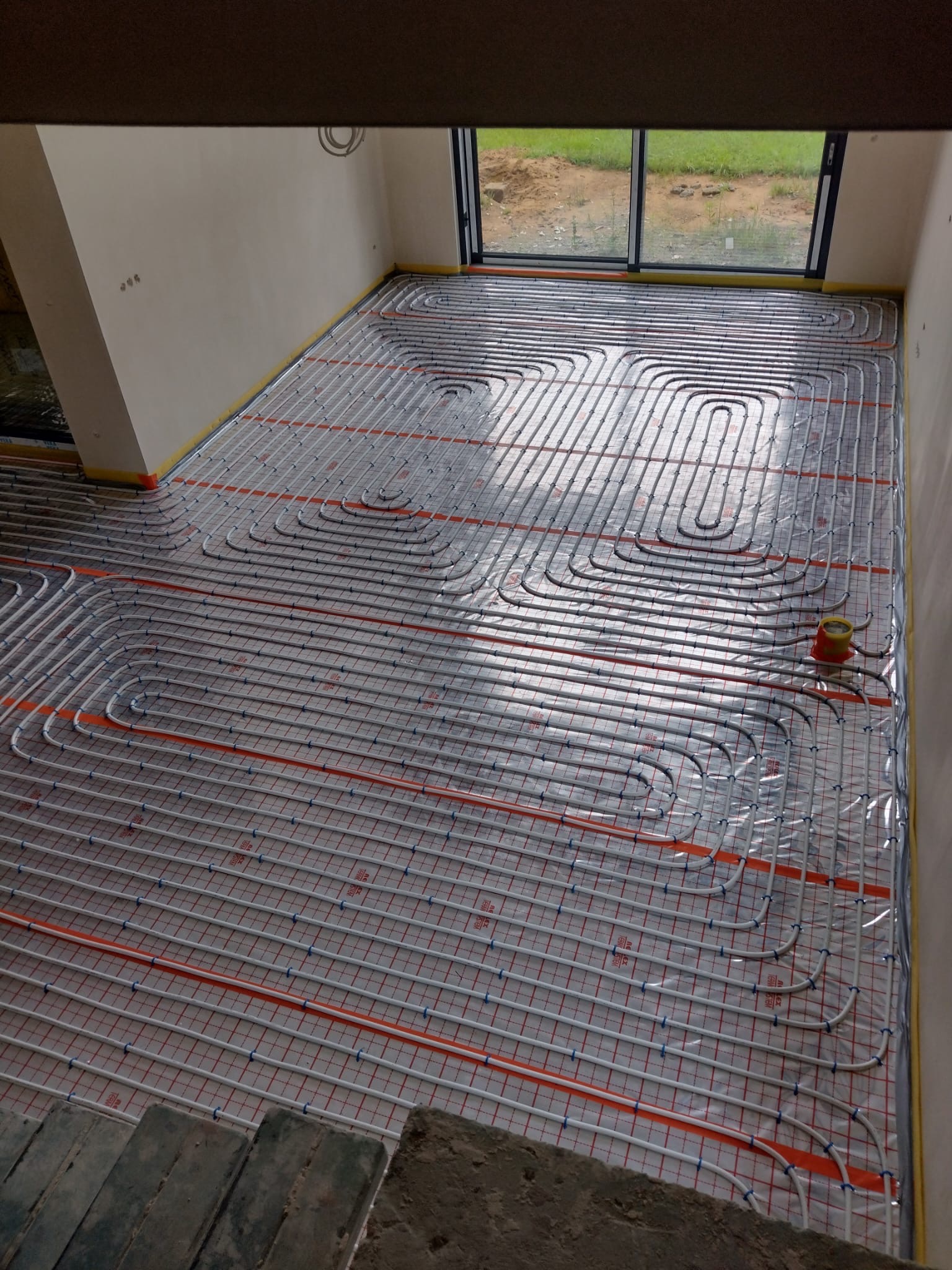 Rozłożone rury ogrzewania podłogowego na folii z siatką, widoczne okno i ściany w surowym stanie, żółta taśma brzegowa przy ścianach, czerwony rozdzielacz w rogu.