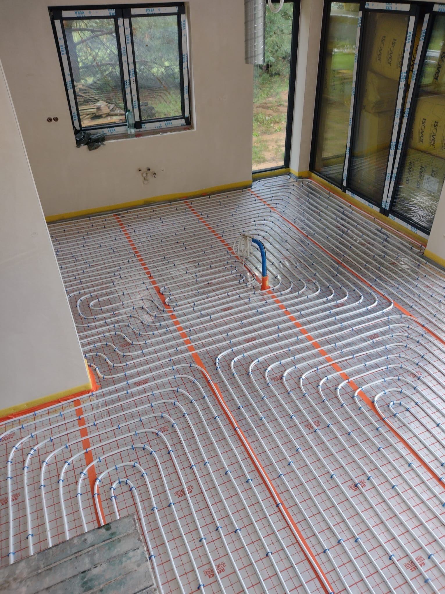 Rozłożona instalacja ogrzewania podłogowego w nowym budynku, widoczne rury grzewcze na folii z siatką, zabezpieczone taśmą żółtą przy ścianach, z fragmentem nieułożonych płyt na dole.