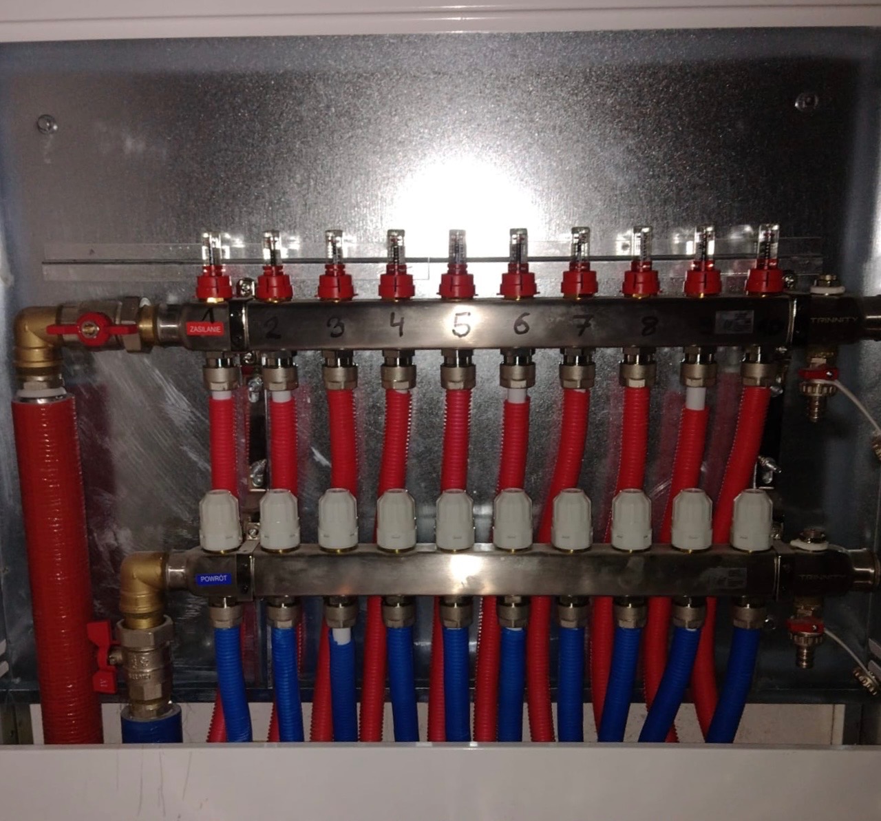 Rozdzielacz ogrzewania podłogowego z rurami czerwonymi i niebieskimi w szafce, widoczne zawory regulacyjne i oznaczenia sekcji.