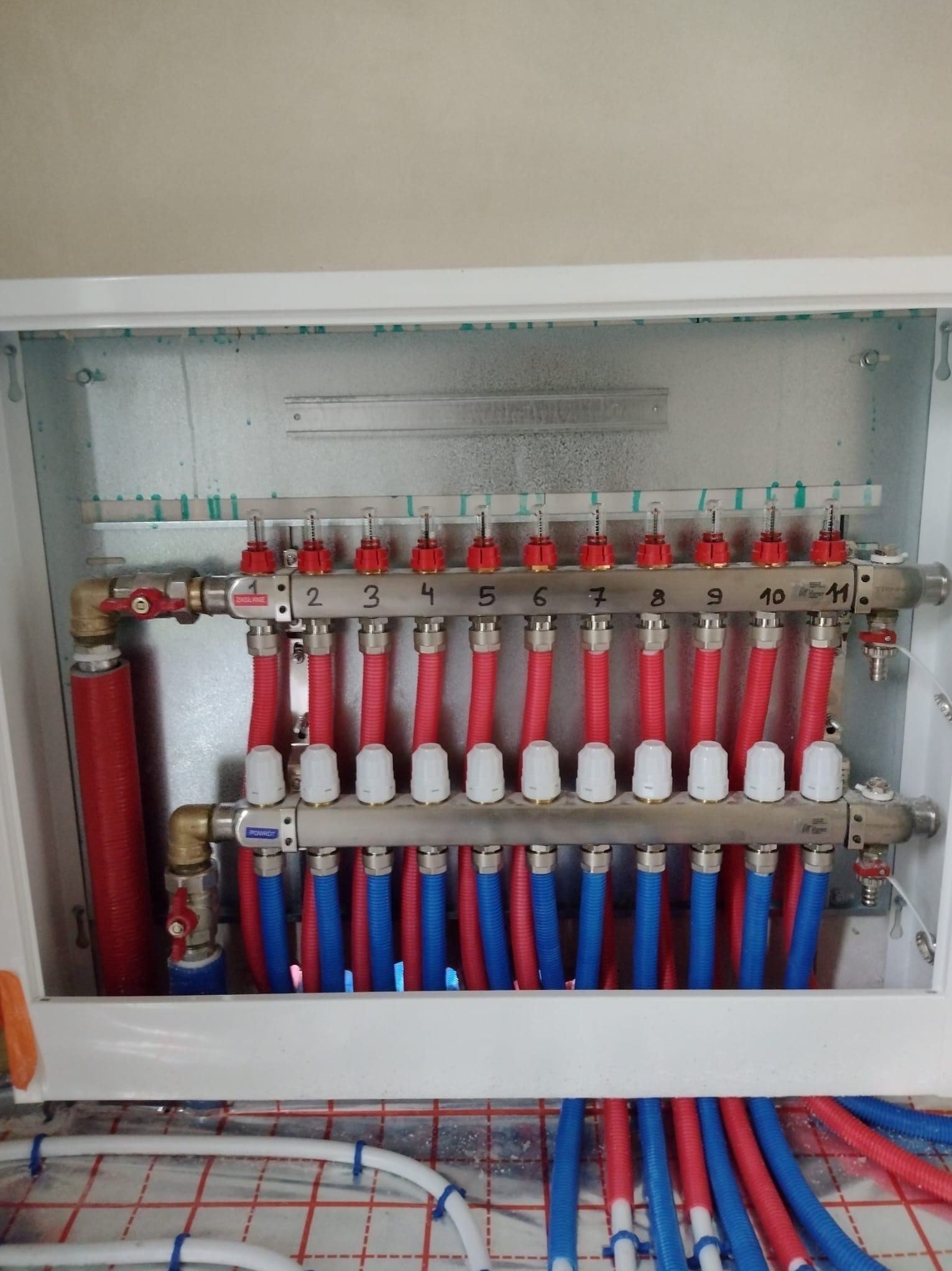 Rozdzielacz ogrzewania podłogowego z numerowanymi sekcjami, widoczne rury w kolorze czerwonym i niebieskim, ułożone na folii z nadrukowaną siatką.