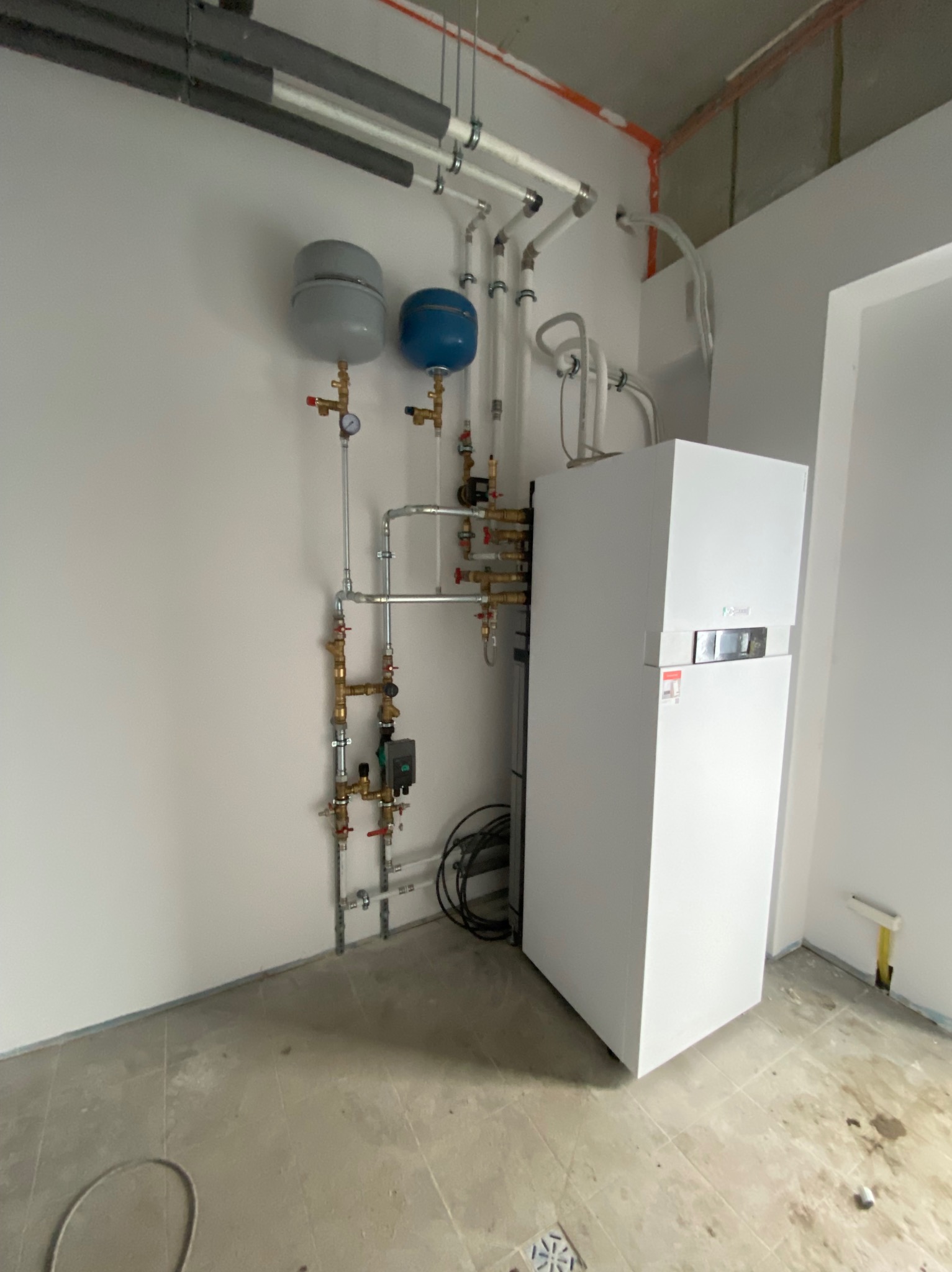 Biały piec grzewczy z podłączonymi rurami, zbiornikami wyrównawczymi i przewodami elektrycznymi w surowym wnętrzu, widoczne elementy instalacji pod sufitem.