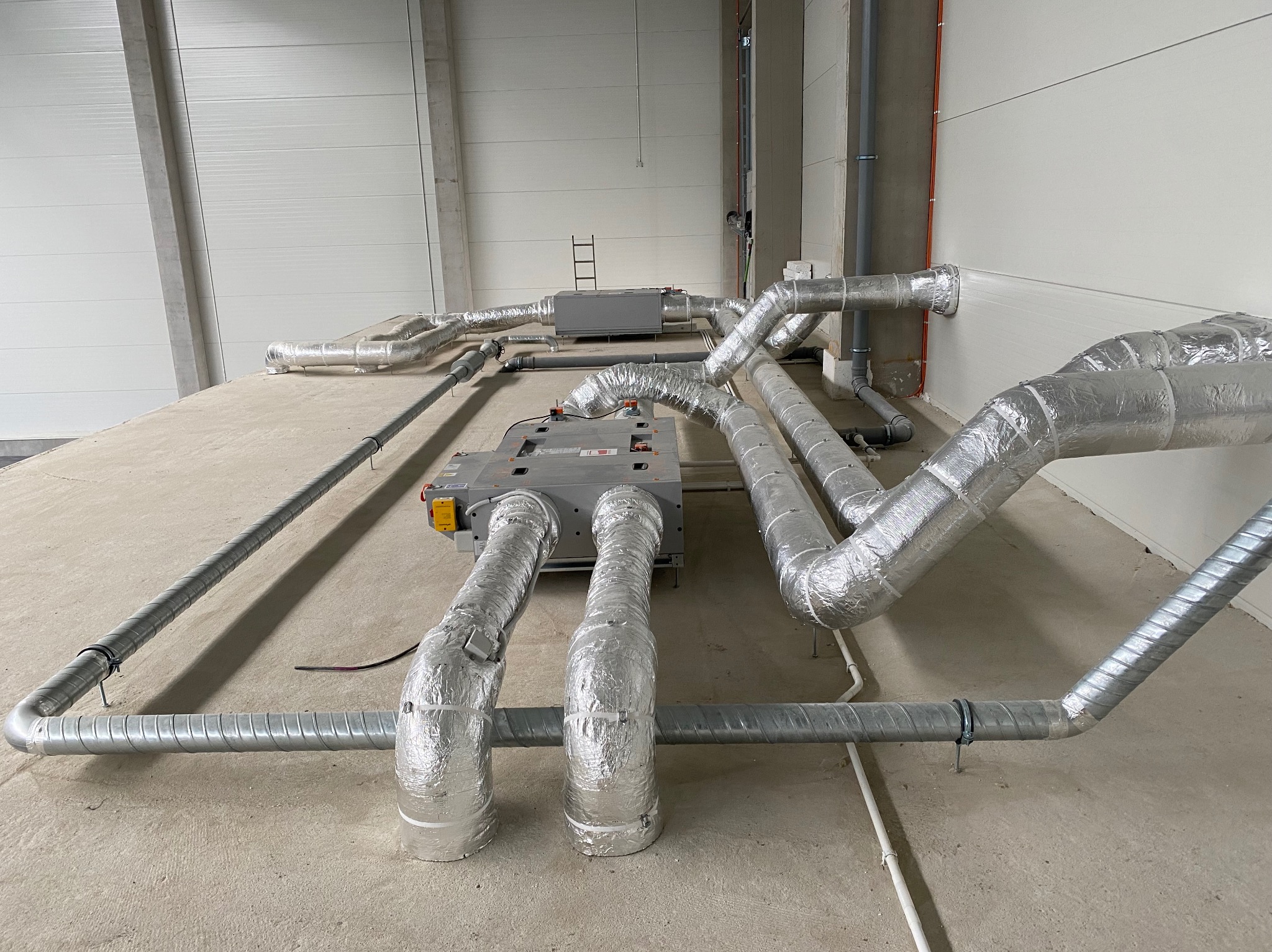 Rozbudowany system kanałów wentylacyjnych z izolacją aluminiową w surowym wnętrzu hali, widoczne dwa centrale wentylacyjne, drabina w tle, szare ściany i betonowa podłoga.