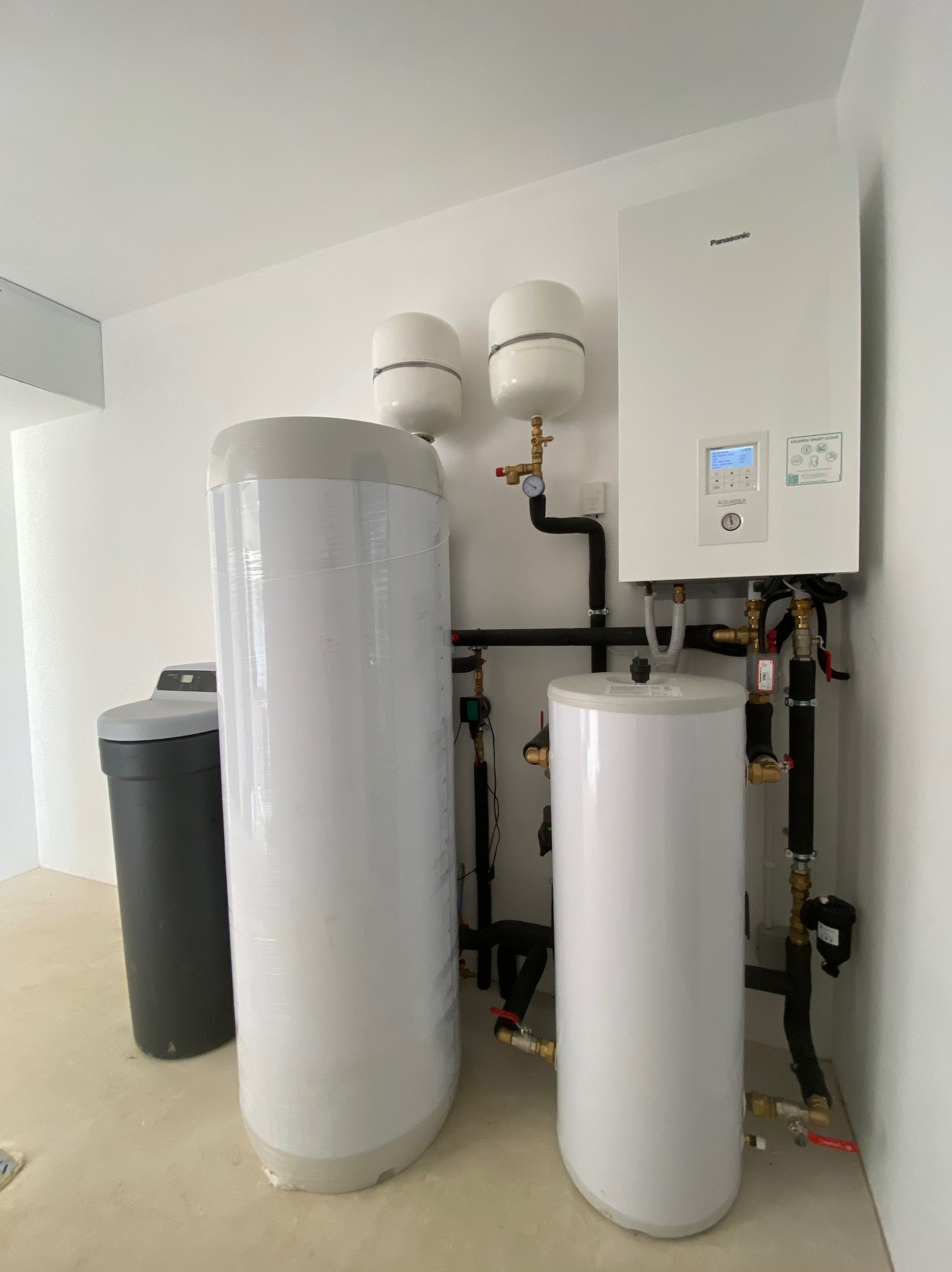 Nowoczesny, biały piec gazowy Panasonic Aquarea Smart Cloud z podłączonymi zbiornikami, rurami i zaworami w minimalistycznym, białym pomieszczeniu; widoczny zmiękczacz wody z boku.