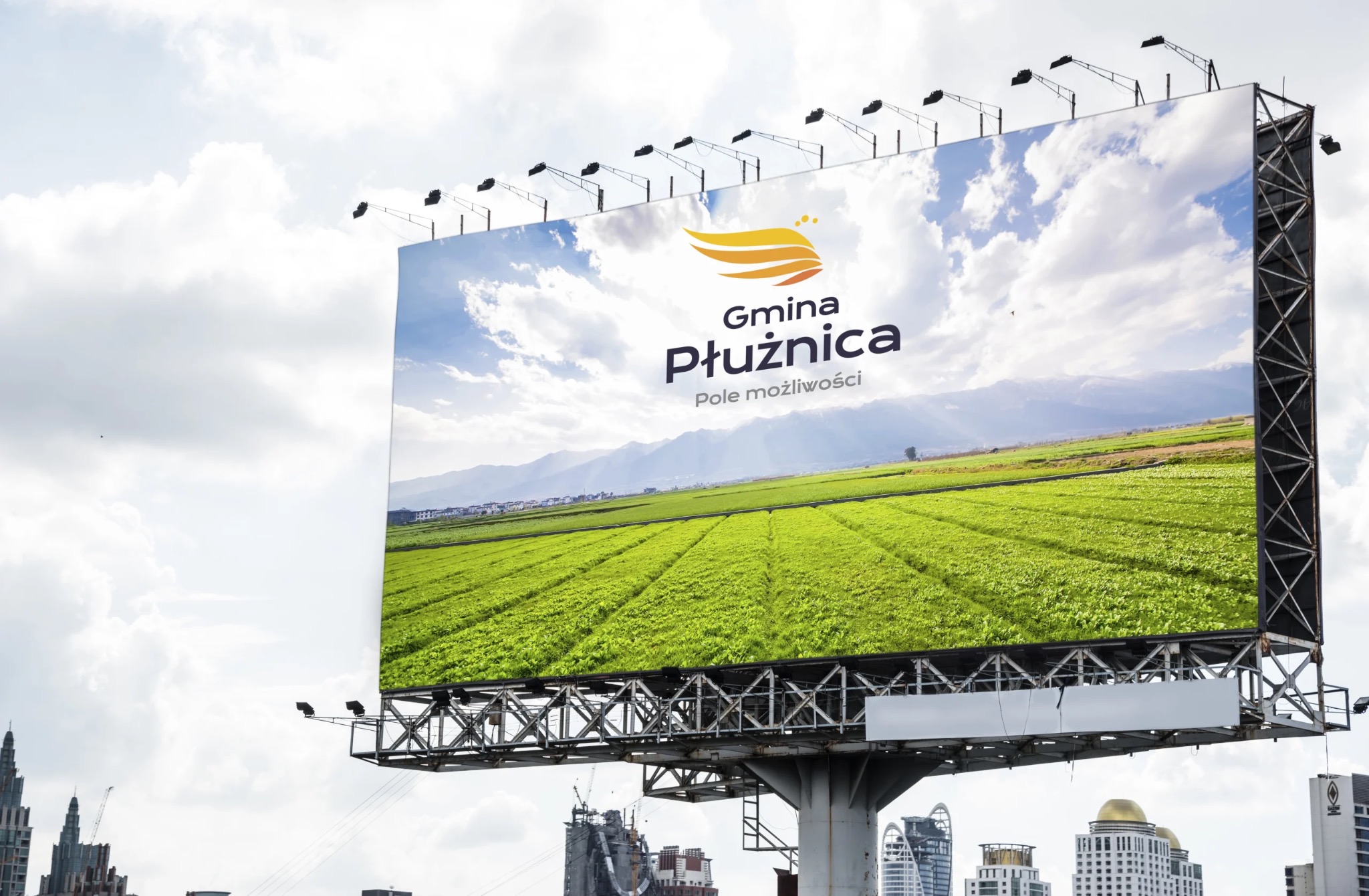 Billboard z krajobrazem gminy Płużnica, na tle miejskiej zabudowy i pochmurnego nieba.