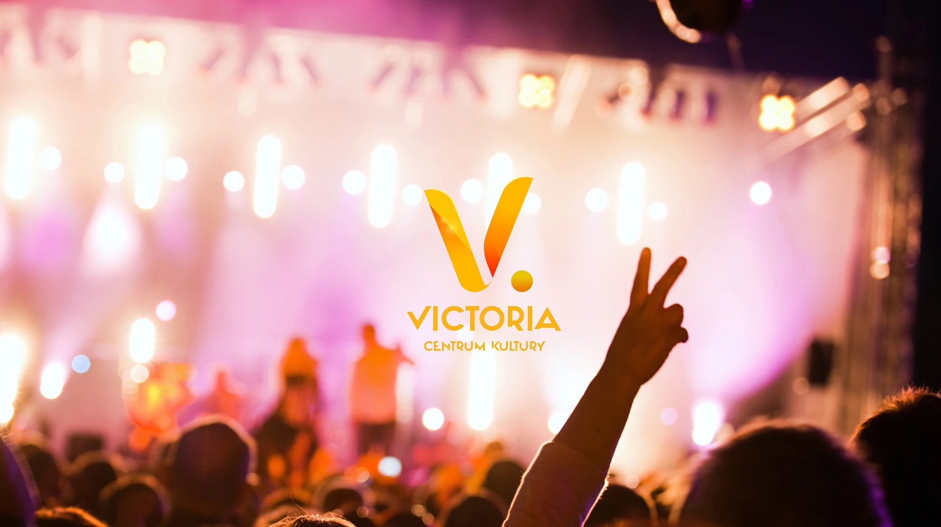 Tłum na koncercie z podniesioną ręką w geście zwycięstwa, scena w tle oświetlona reflektorami i logo 'Victoria Centrum Kultury' w centralnej części kadru.